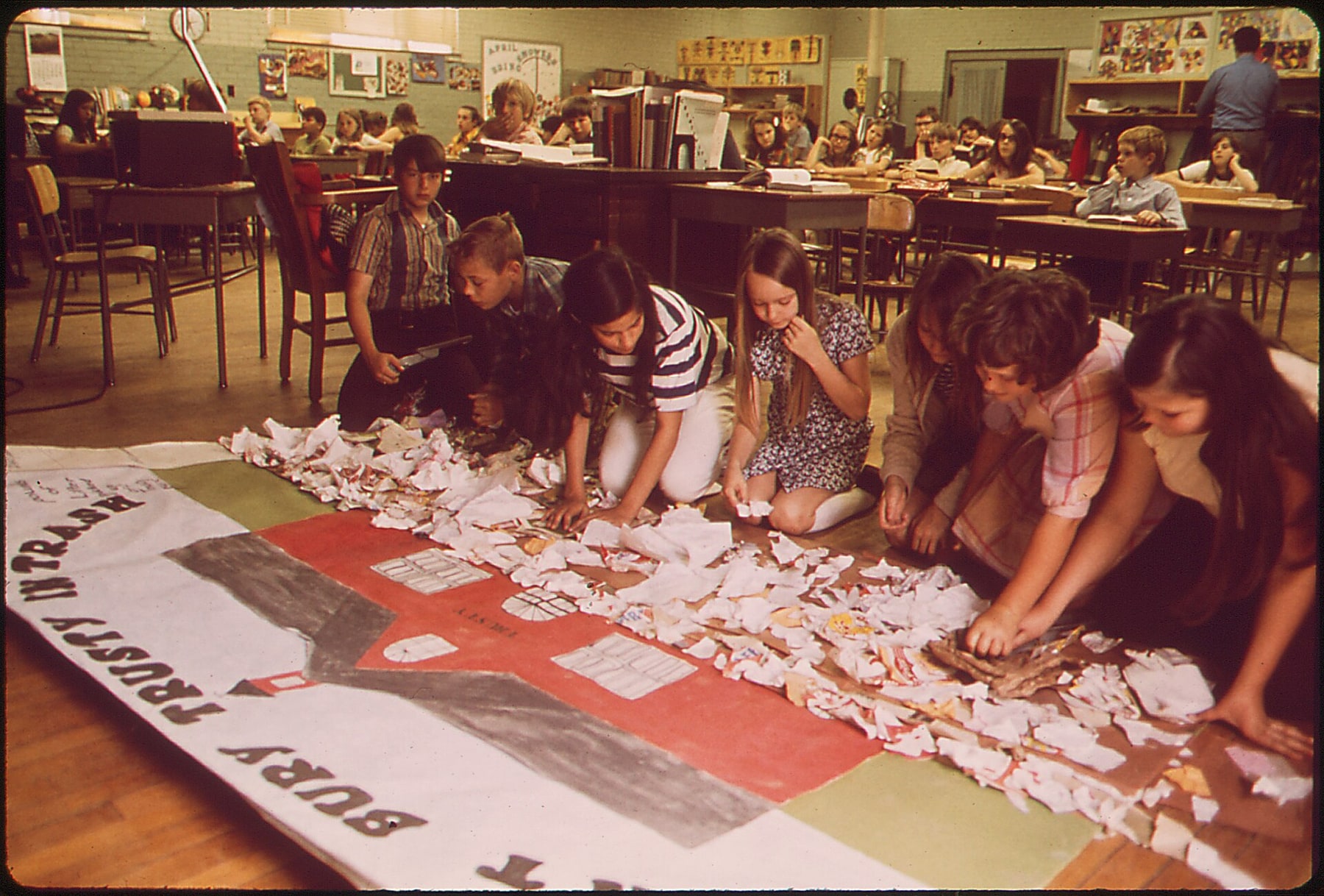 Students kneel around a large handmade ecology display while classmates work at desks behind them.