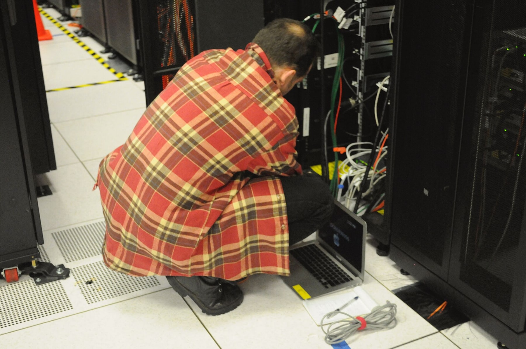 A technician works on an open server rack with a laptop in a data center.