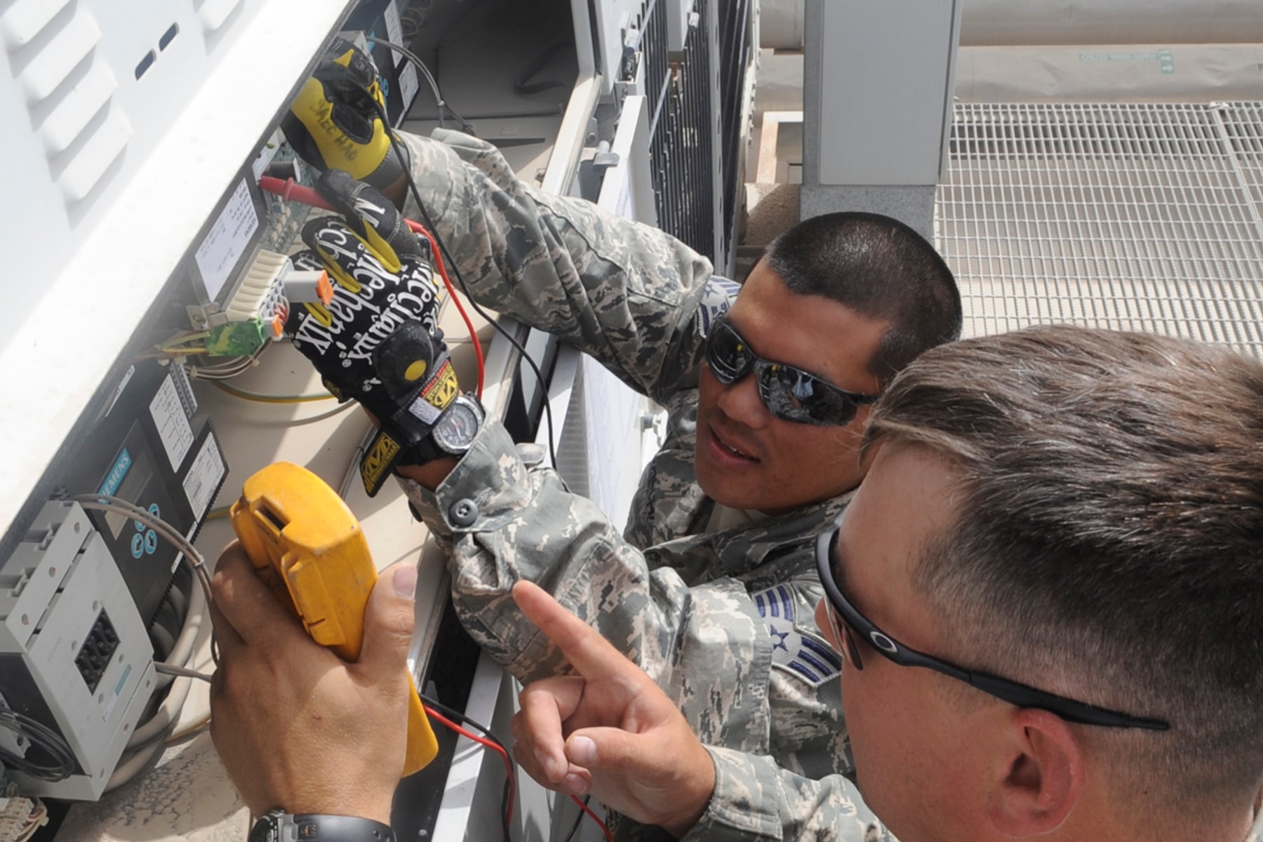 Two technicians test voltage connections inside a large outdoor cooling unit.