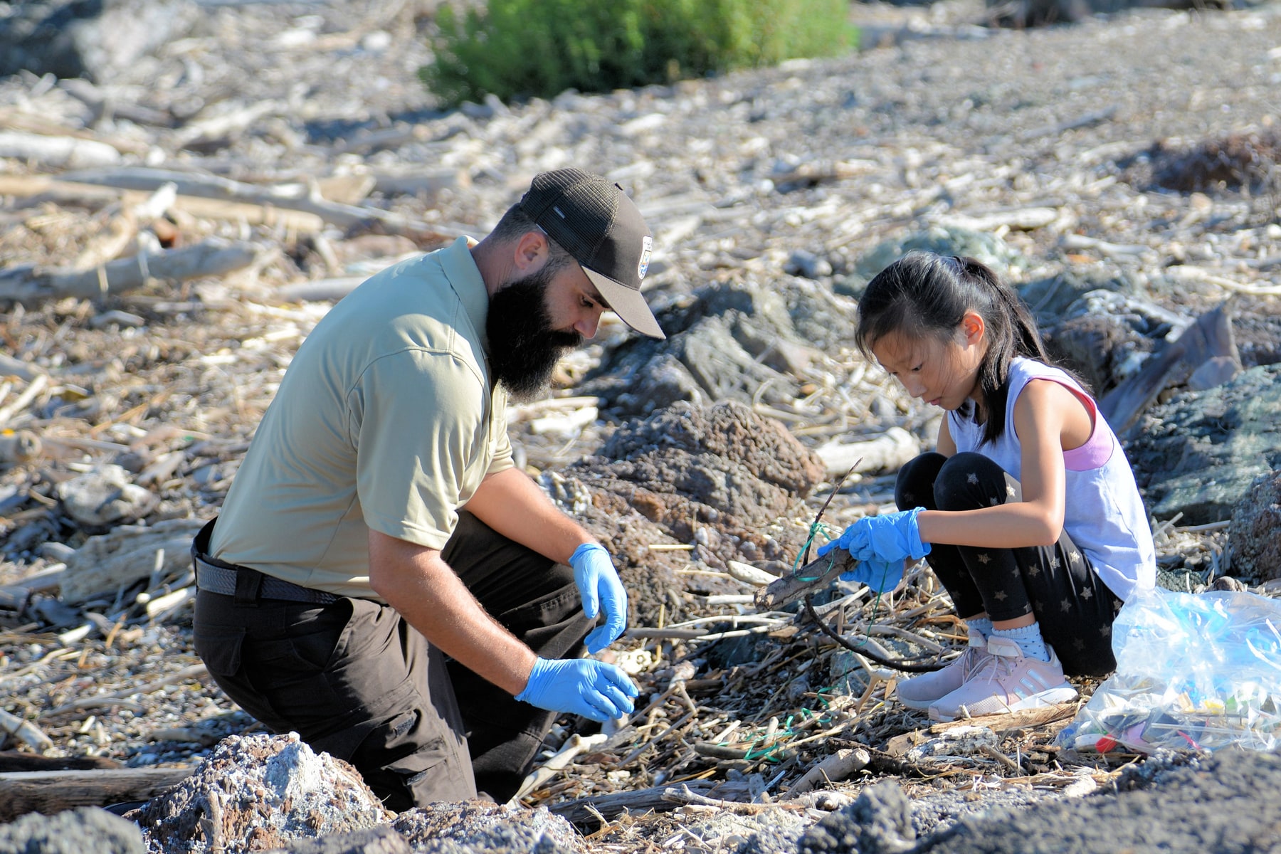A young person and a refuge worker collect plastic debris together on a rocky shoreline.