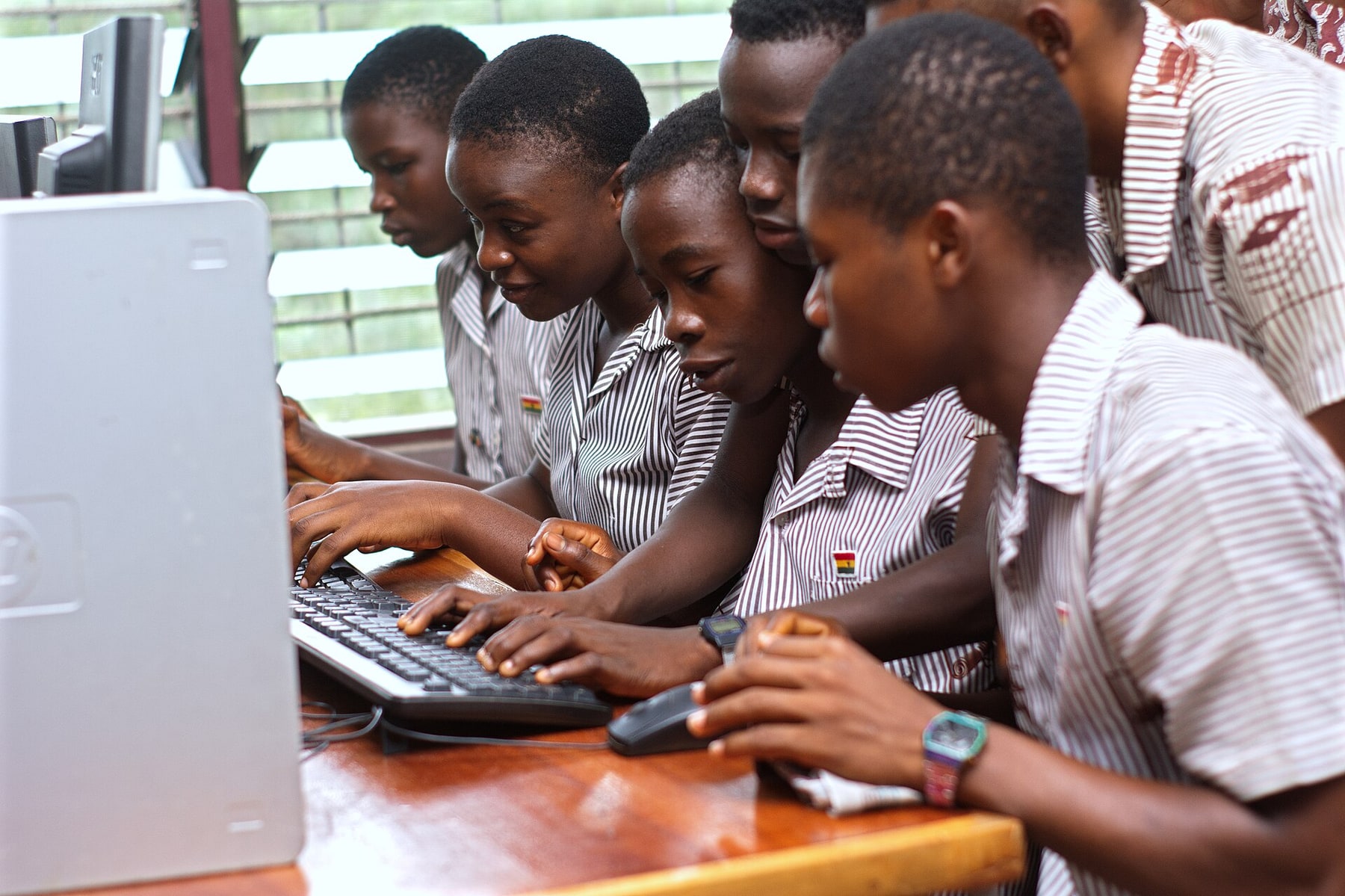 Students gather around a classroom computer while two students use the keyboard and mouse.