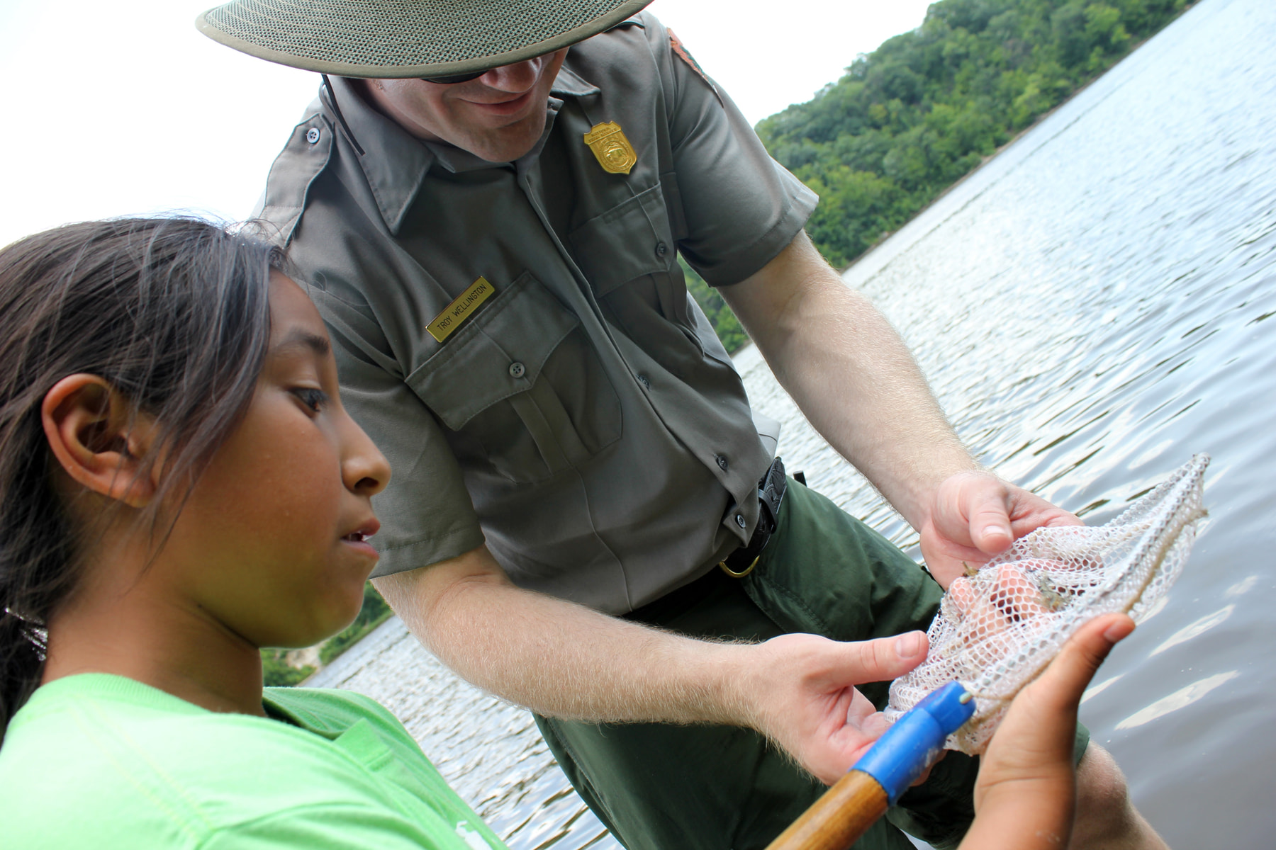 A student and park ranger examine a small aquatic animal beside a lake.