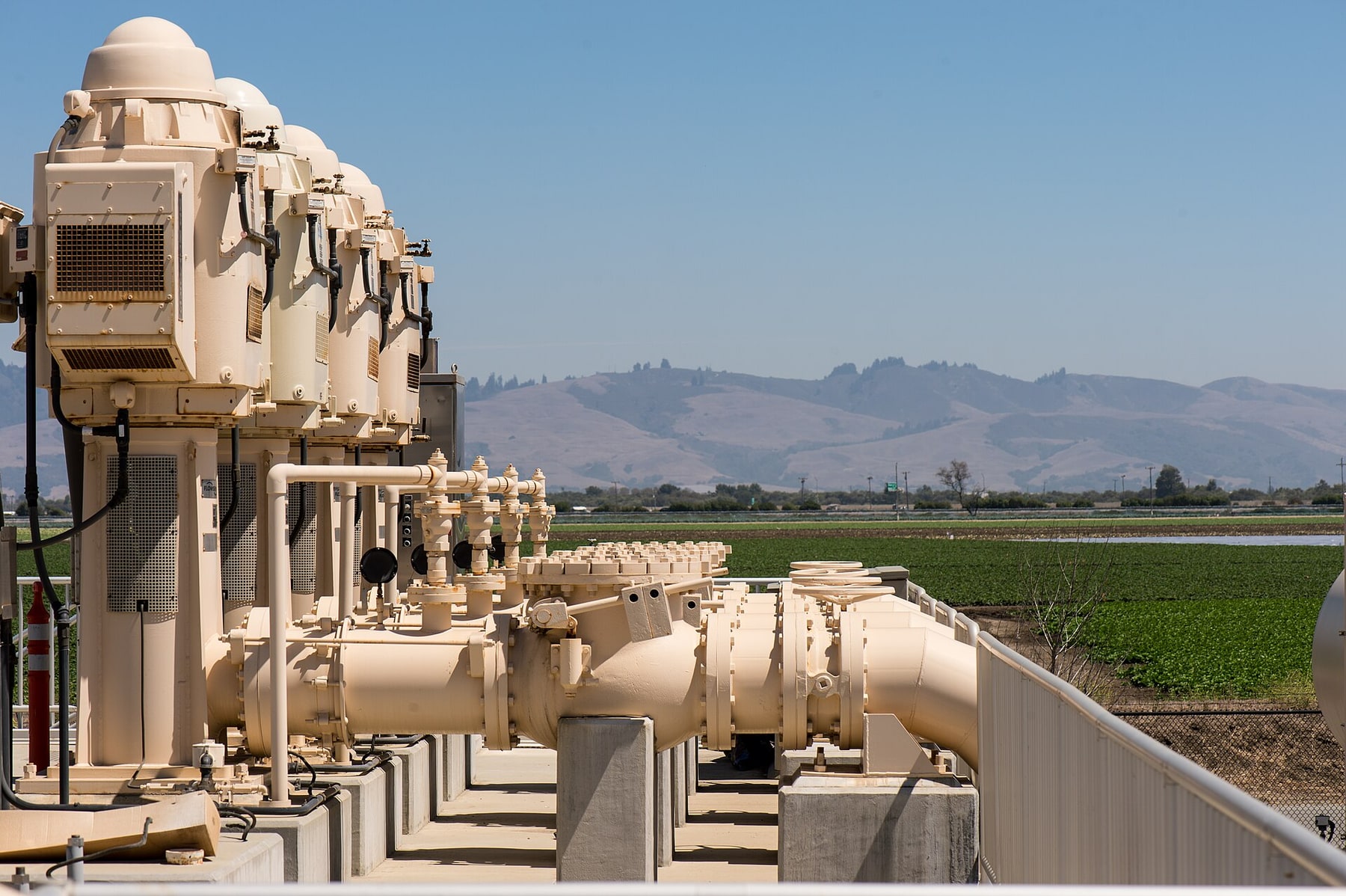 A row of large beige pumps and connected pipes stands outdoors beside farm fields and low hills.